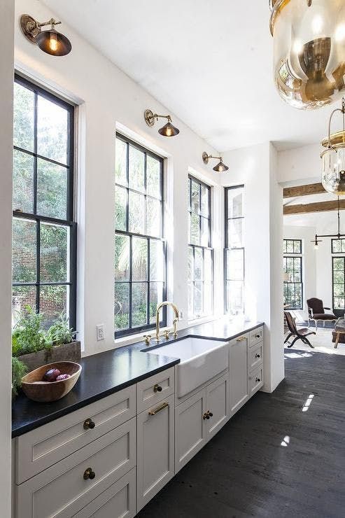 una cucina lineare bianca vintage con ripiani neri, ampie finestre, applique in ottone ed eleganti lampade a sospensione