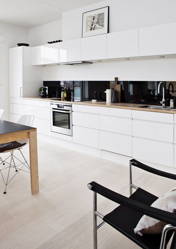 una cucina bianca minimalista in linea con ripiani biondi in macellaio, un alzatina in vetro nero e una zona pranzo