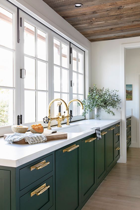 una cucina moderna verde cacciatore con ripiani in pietra bianca, maniglie e infissi dorati e una finestra invece di un alzatina