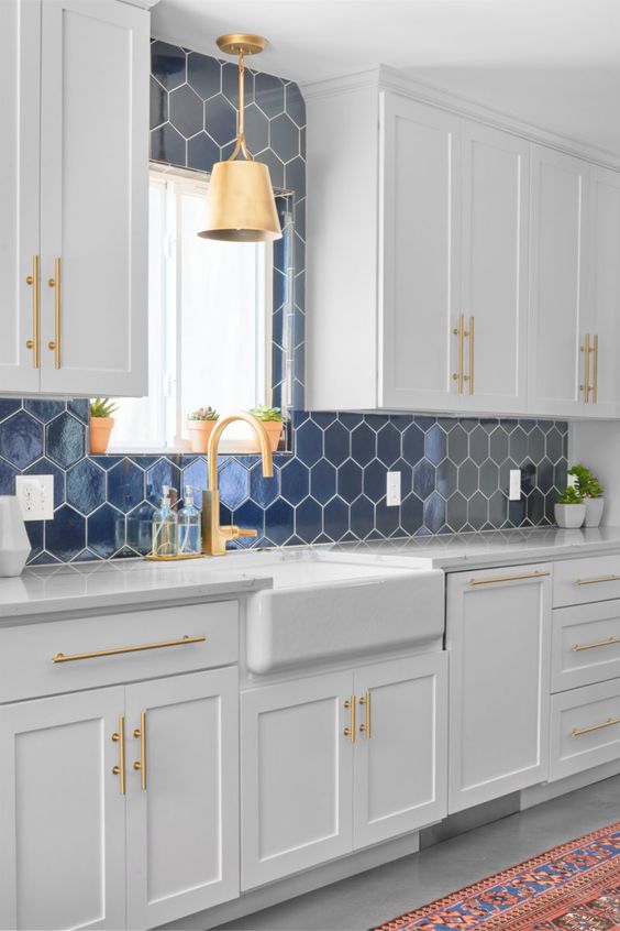 un'elegante cucina bianca con un backsplash in piastrelle esagonali blu scuro e malta bianca, infissi dorati e una lampada a sospensione è sorprendente