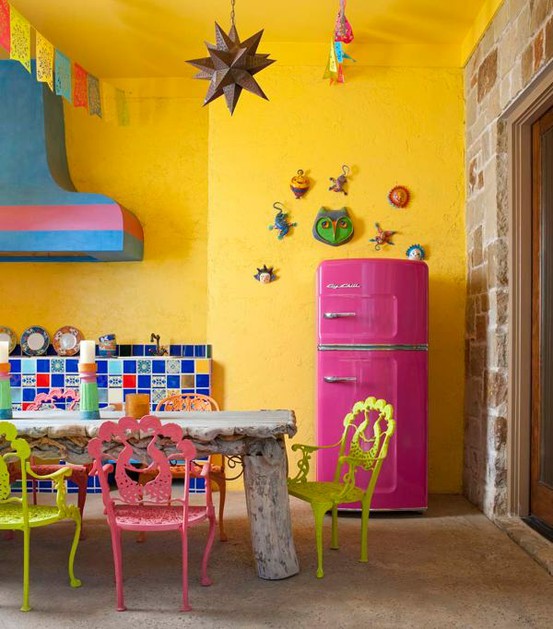 uno spazio colorato con pareti gialle, un alzatina in piastrelle colorate, un tavolo in legno grezzo imbiancato a calce, sedie gialle e rosa e decorazioni colorate più un frigorifero rosa