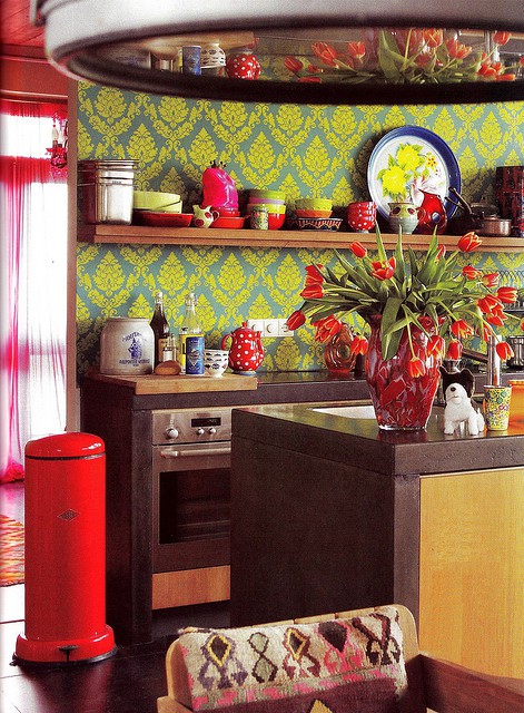 una cucina colorata con tocchi boho: mobili marroni, carta da parati stampata, tocchi di rosso e stampe boho sembrano insolite
