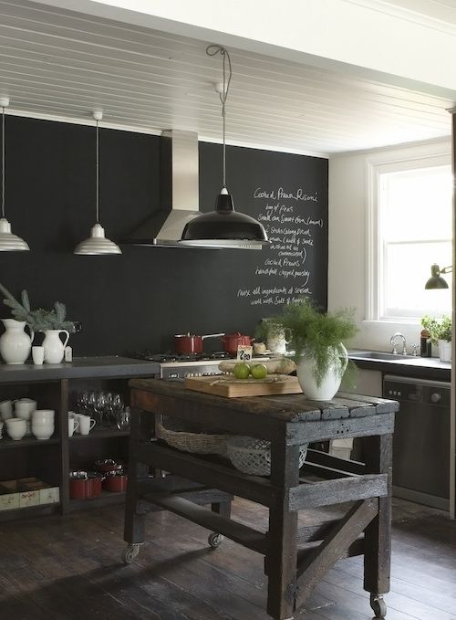 una cucina scura con una parete lavagna, isola cucina in legno, armadi in cemento e pietra e lampade a sospensione