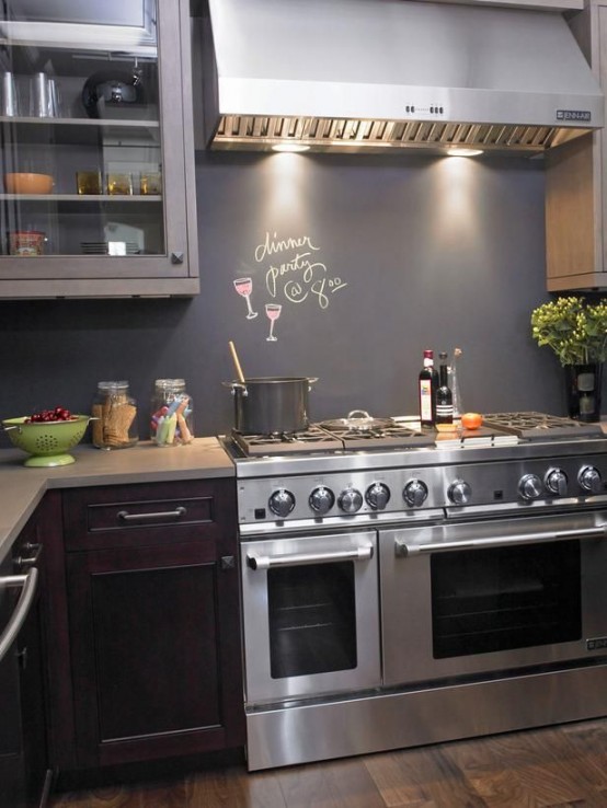una cucina scura con mobili inferiori viola e superiori grigi, un alzatina a lavagna e luci sopra il fornello è molto chic