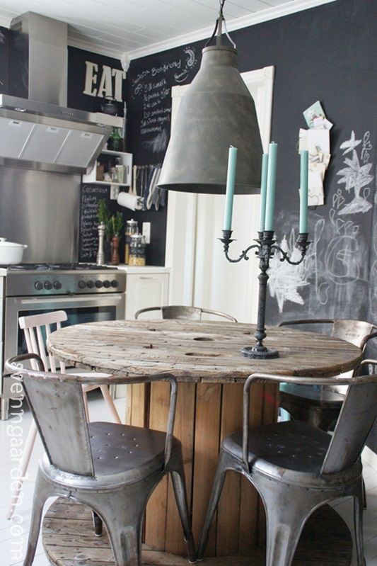 una cucina nordica con pareti a lavagna, elettrodomestici e sgabelli in acciaio inossidabile, una lampada di cemento e un tavolo di legno