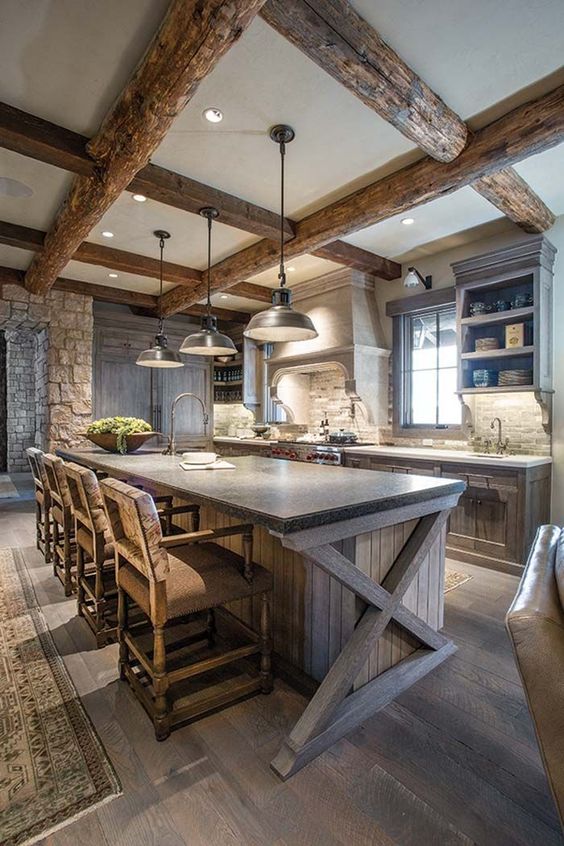 una cucina da chalet con mobili in legno grezzo e isola della cucina, ripiani in pietra, travi in ​​legno e lampade a sospensione e sgabelli vintage