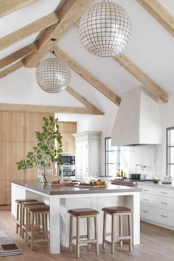 una moderna cucina rustica neutra con ante bianche e isola cucina, alzatina in marmo, travi in ​​legno chiaro e lampade a sfera