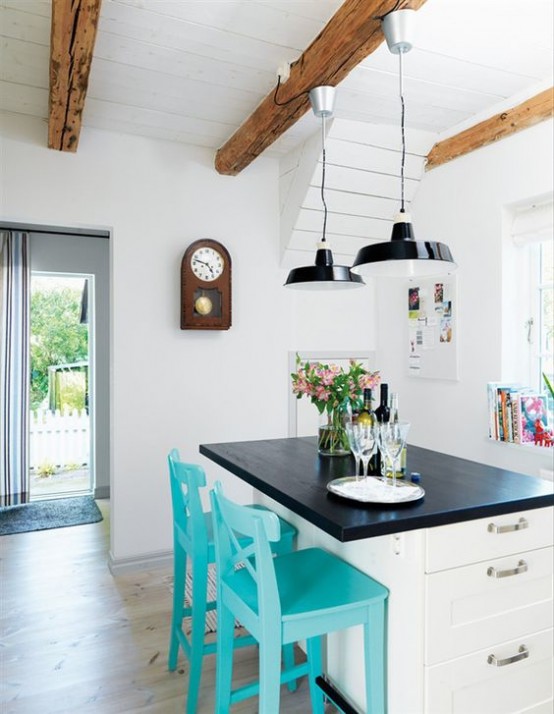una cucina bianca da fattoria con accoglienti mobili bianchi, lampade a sospensione nere e travi in ​​legno che aggiungono interesse e calore allo spazio pulito e arioso