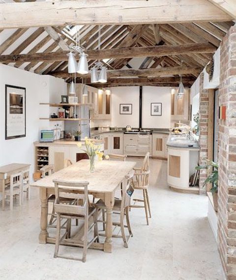 una moderna cucina naturale con eleganti mobili bianchi, travi in ​​legno grezzo e lampade a sospensione, un set da pranzo in legno e sedie è molto creativa