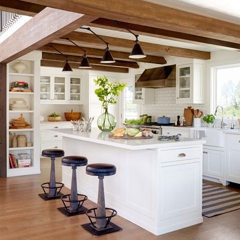 una moderna cucina rustica neutra con mobili bianchi, travi in ​​legno sul soffitto con applique nere, sgabelli neri e vegetazione per un tocco di freschezza