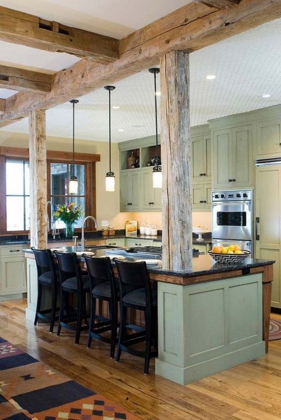una cucina colonica con mobili verde oliva, controsoffitti grigi, travi in ​​legno sul soffitto e pilastri più lampade a sospensione