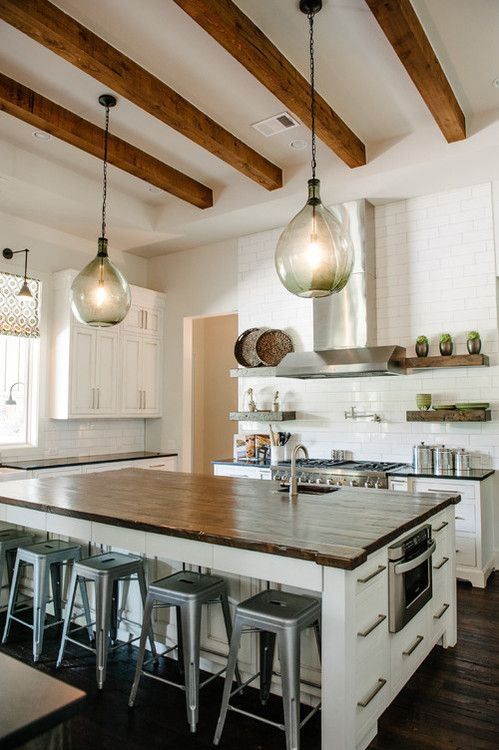 una cucina bianca con eleganti mobili da fattoria, una grande isola della cucina, lampade a sospensione, travi in ​​legno dai toni ricchi che aggiungono intimità allo spazio