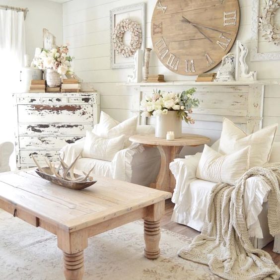un soggiorno shabby chic neutro con pareti bianche, una credenza shabby, un orologio in legno e un tavolo e alcuni tessuti