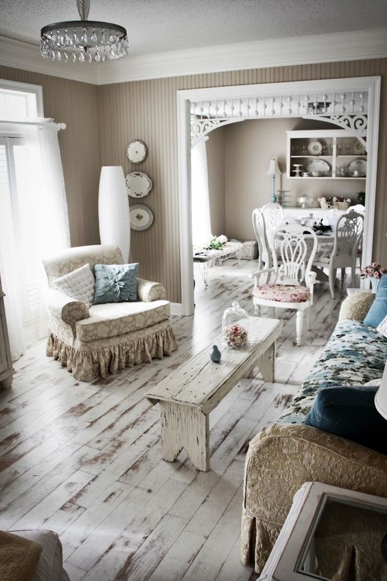 un soggiorno shabby chic con un divano floreale blu, sedie stampate e una panca bianca, un lampadario di cristallo e cuscini pastello