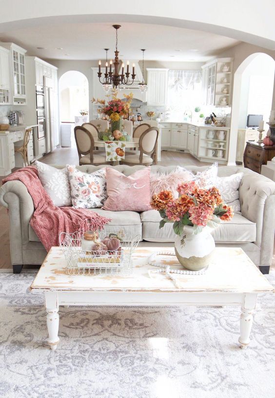 un soggiorno shabby chic con un divano grigio, un tavolo shabby bianco, cuscini rosa, coperte e fiori