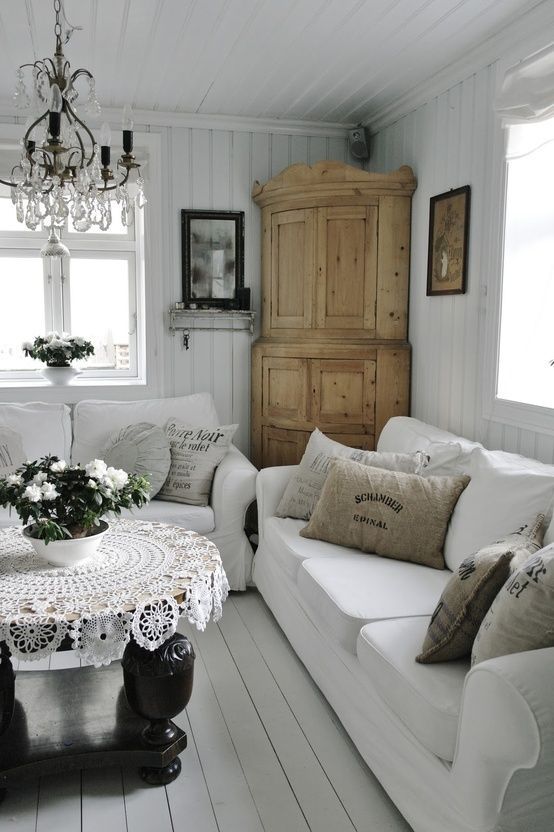 un soggiorno dal bianco vintage allo shabby chic con divani bianchi, un armadio in legno, un lampadario di cristallo, un tavolo nero, una tovaglia all'uncinetto e fiori