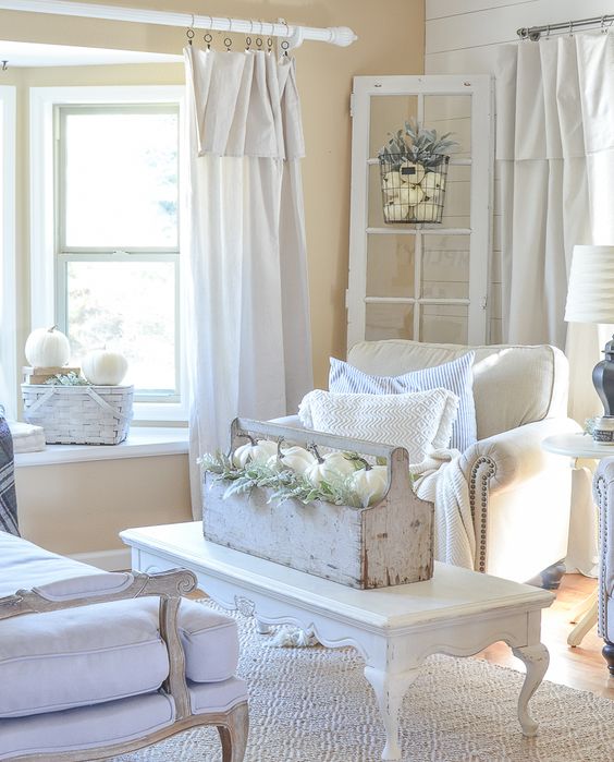 uno spazio bianco shabby chic con mobili eleganti, un tavolino basso, una cassetta degli attrezzi con fiori e zucche bianche