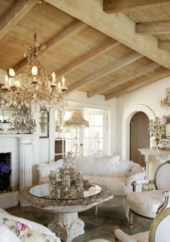 un raffinato soggiorno shabby chic francese con travi in ​​legno, camino, mobili eleganti e chic, un lampadario di cristallo e uno splendido tavolo in pietra