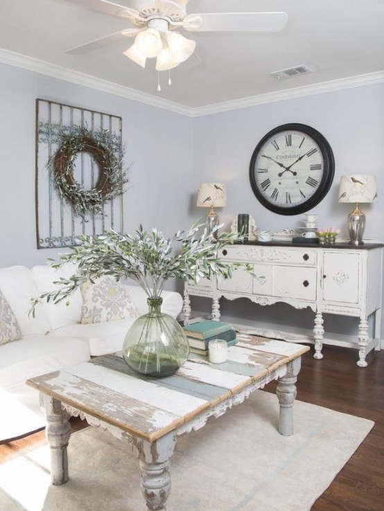 un soggiorno shabby chic con pareti blu pastello, raffinati mobili neutri, una ghirlanda e un orologio, un tavolo basso e lenzuola neutre