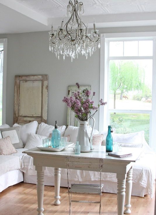 un soggiorno shabby chic bianco con mobili vintage, un lampadario di cristallo, vasi blu, fiori e uno specchio