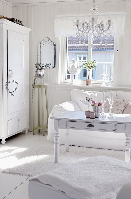 un soggiorno shabby chic bianco con mobili imbiancati, un lampadario di cristallo, vegetazione in vaso e piante e cuscini floreali
