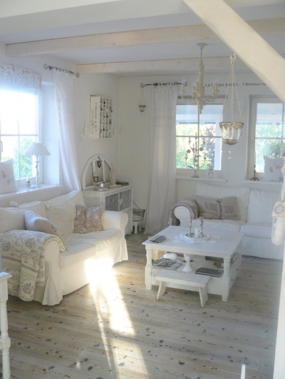 un soggiorno shabby chic bianco con mobili vintage, travi in ​​legno, un lampadario di cristallo, pizzo e tende bianche