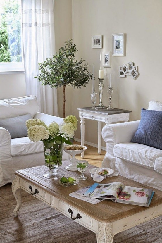 un raffinato soggiorno shabby chic in colori neutri, con mobili bianchi, un tavolo basso, cuscini blu e vegetazione in vaso