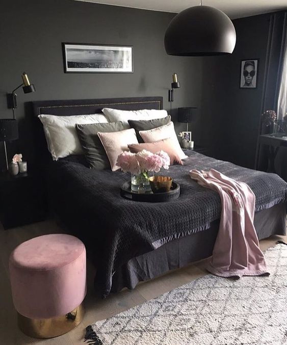 una camera da letto lunatica con pareti grigie, un letto nero, comodino e una lampada a sospensione, biancheria da letto rosa e nera e un ottomano rosa