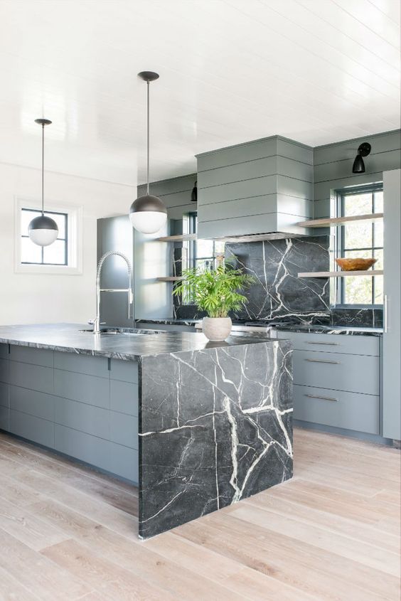 una cucina in legno grigio con alzatina in marmo nero e un'isola abbinata con un piano di lavoro a cascata