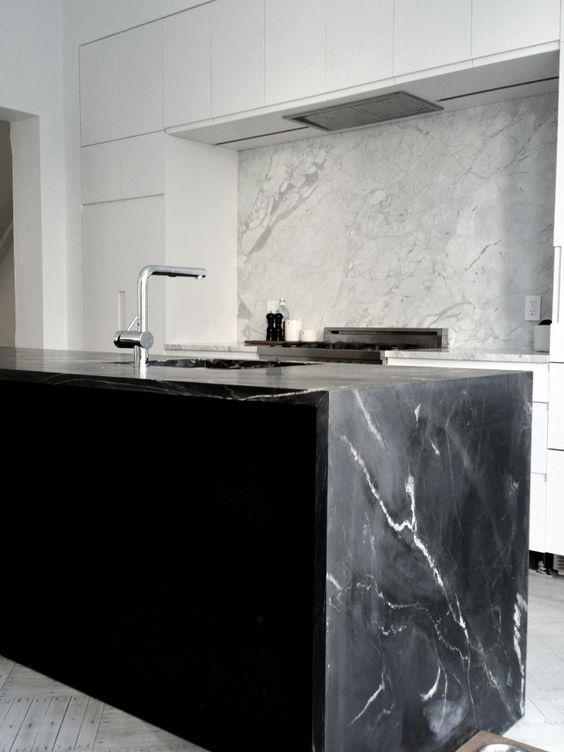 una cucina bianca minimalista e un'isola da cucina nera con un bel ripiano in marmo nero che riecheggia con un backsplash in marmo bianco