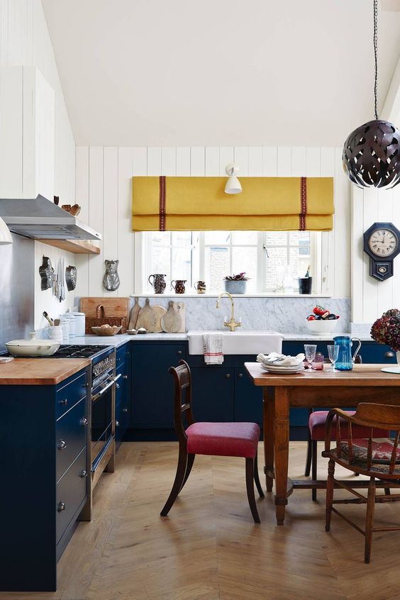 una cucina blu scuro e bianca con ripiani in pietra bianca, una tenda gialla e sedie rosse luminose sembra fresca ed eclettica