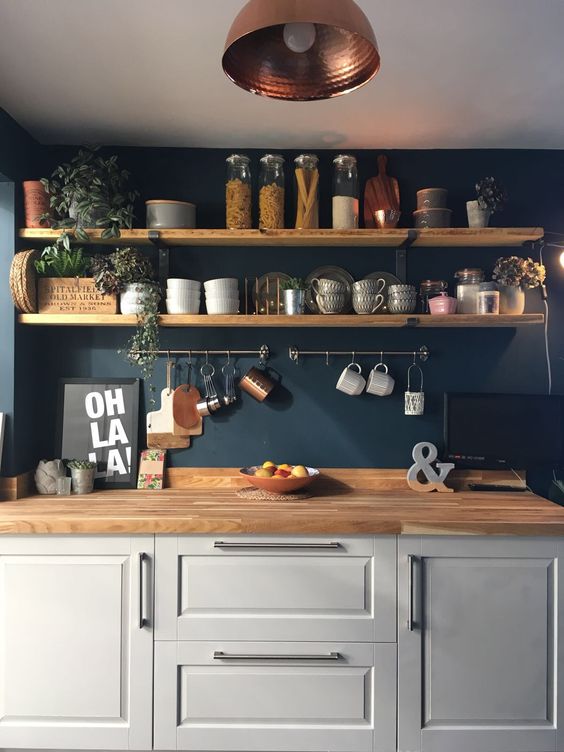 una cucina grigio tortora con un muro blu scuro, mensole in legno e ripiani da macellaio più tocchi di rame e verde