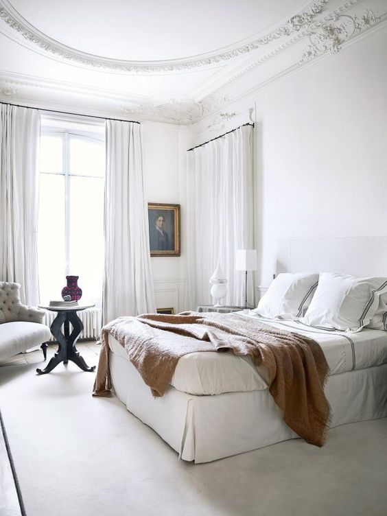 una camera da letto parigina bianca con tende ariose, un letto imbottito, un'opera d'arte e un'elegante sedia trapuntata nell'angolo