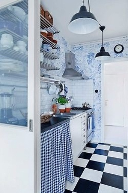 una cucina retrò con pavimento a quadri, carta da parati floreale blu e bianca, scaffali aperti e lampade a sospensione