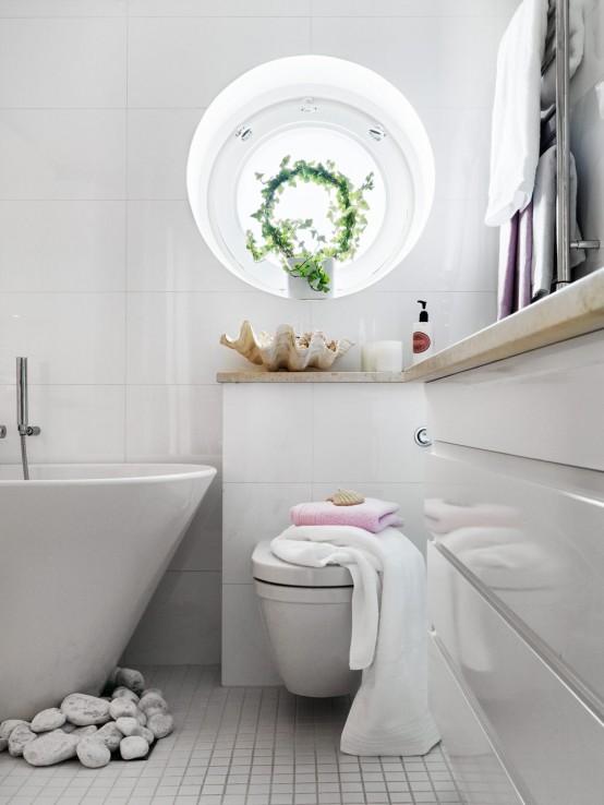 un bagno bianco minimalista con un'elegante vanità con un ripiano in pietra neutra, una vasca ovale con ciottoli, una finestra rotonda e vegetazione
