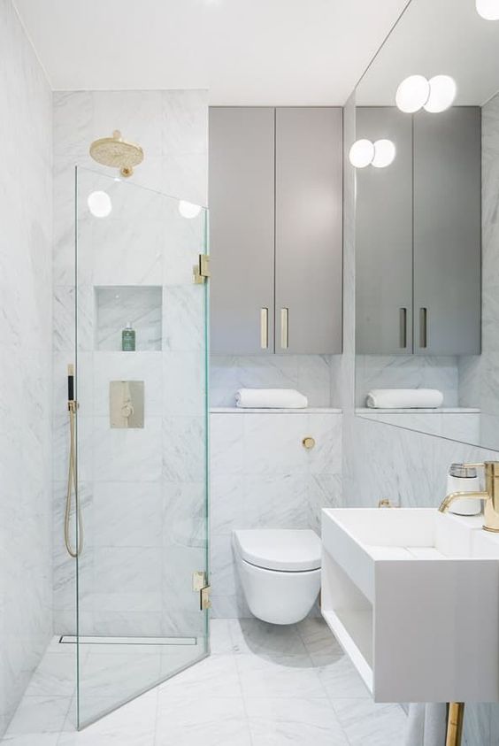 un minuscolo bagno minimalista con piastrelle in marmo bianco, eleganti armadi grigi, tocchi dorati e uno spazio doccia triangolare