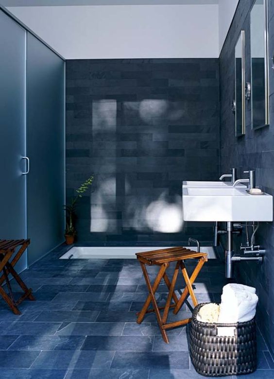 un bagno blu scuro e grigio con una vasca incassata bianca e lavandini più sgabelli in legno piegati e un cesto per la conservazione