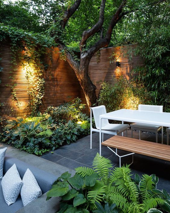 un giardino in una residenza di città contemporanea con piastrelle in pietra, mobili minimalisti in legno e metallo, una vegetazione lussureggiante e un albero che cresce