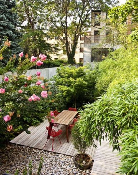 un piccolo giardino in una casa a schiera con un ponte di legno, ciottoli, vegetazione in crescita e alberi più sedie rosse e un tavolo da pranzo
