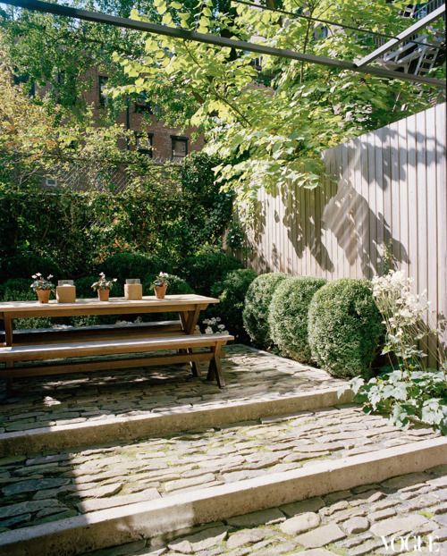 un giardino di una casa a schiera con gradini, vegetazione e bosso in crescita e un semplice set da pranzo in legno con panchine