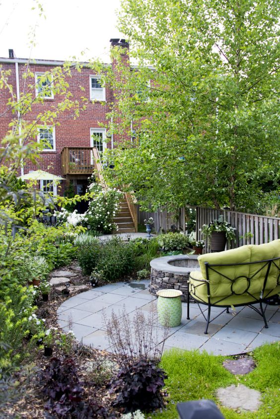 un elegante giardino in una casa a schiera con una piattaforma in pietra, un pozzo del fuoco, un divano verde, fiori piantati e vegetazione