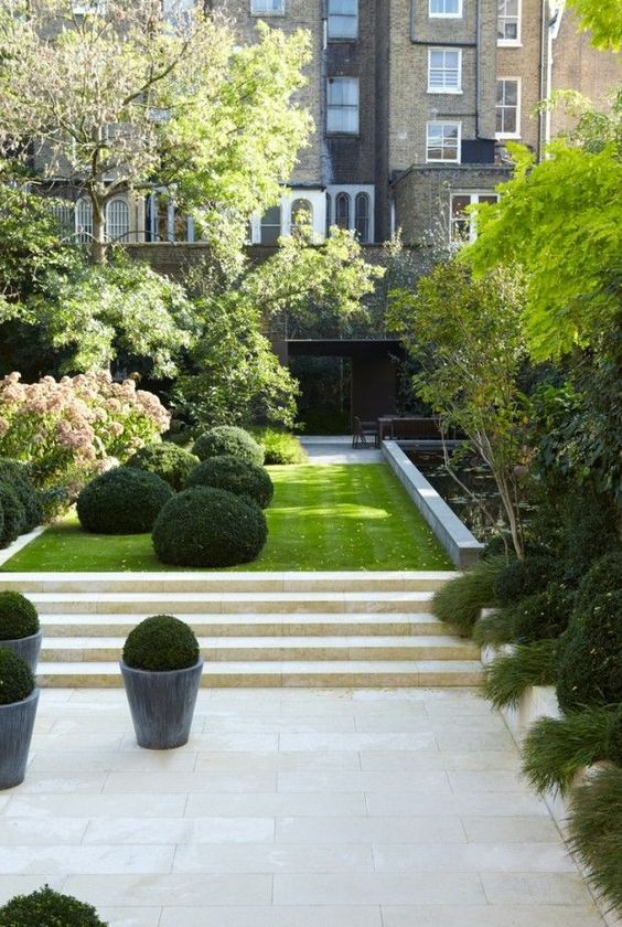 un elegante giardino minimalista in una casa a schiera con piastrelle e gradini in pietra, un prato, bosso, alberi e arbusti più un'aiuola