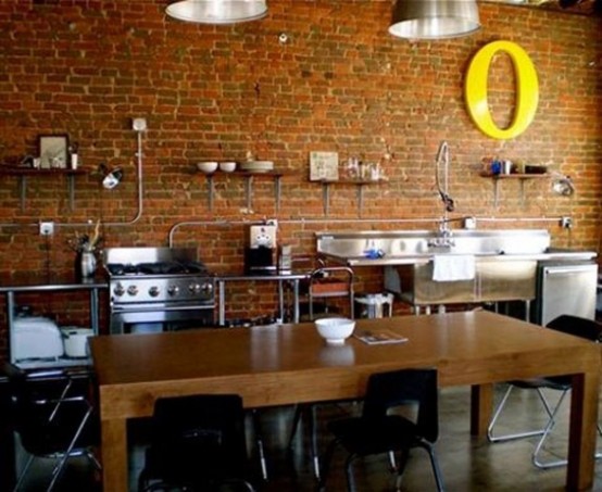 una cucina completamente industriale con un muro di mattoni rossi e superfici metalliche lucide, una lettera gialla in grassetto e un tavolo di legno