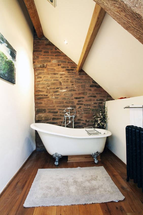 un bagno in mansarda di ispirazione vintage con un muro in pietra, travi in ​​legno, una vasca con i piedini e un radiatore nero
