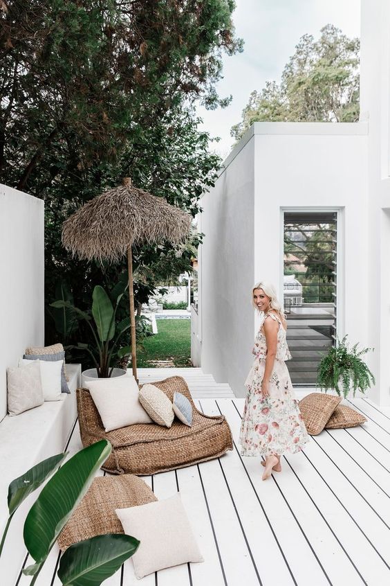 un portico tropicale con un pavimento in legno imbiancato, mobili di tela da imballaggio, un ombrello, piante in vaso e cuscini