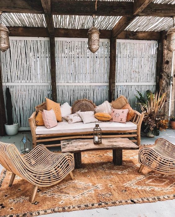 un patio tropicale boho con una panca di legno e molti cuscini, sedie in rattan accattivanti, un tavolo in legno e un tappeto boho più lanterne marocchine
