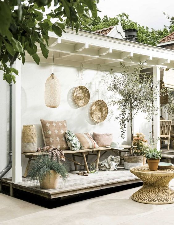 un accogliente patio tropicale boho con un tavolo in rattan, una panca in legno con molti cuscini, fioriere in vimini e cesti decorativi