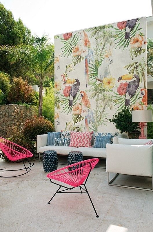 un colorato patio tropicale con un divano bianco e cuscini colorati, sedie rosa acceso, un luminoso muro ad acquerello e piccoli sgabelli