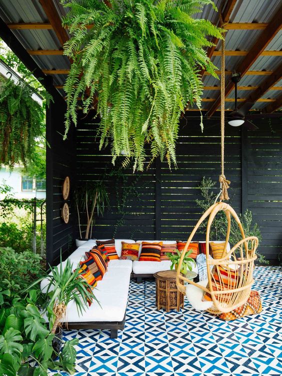 un bellissimo patio tropicale con un tetto e vegetazione che pende, con un divano a forma di L con cuscini colorati, una sedia in rattan e un pavimento di piastrelle a mosaico