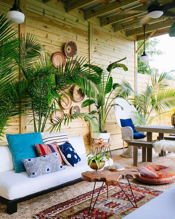 un patio tropicale boho con piante in vaso, un divano bianco, tappeti boho e un pouf, un tavolo vivente e cesti decorativi sul muro